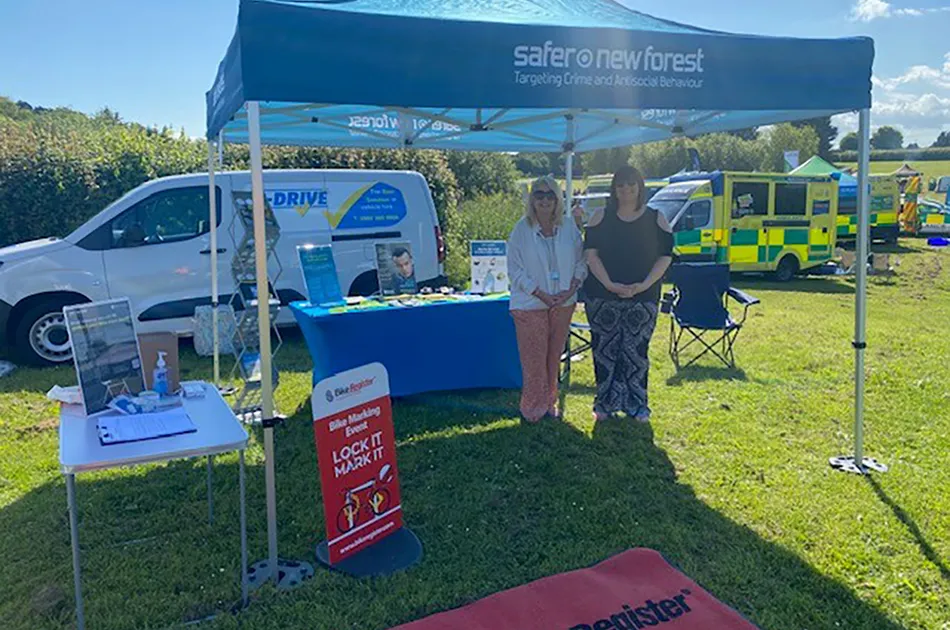 Two women standing under a blue 'Safer New Forest' gazebo at an outdoor community event, with information tables, a van, and emergency vehicles in the background on a sunny day.
