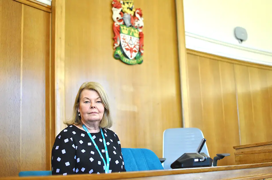 A woman with blonde hair sits in a courtroom on a blue chair, wearing a black and white patterned top and a lanyard. A crest is mounted on the wood-panelled wall behind her.