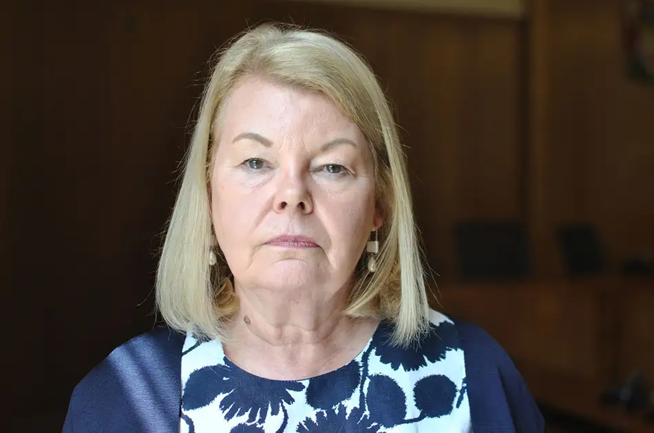 A woman with straight blonde hair and light skin looks directly at the camera with a neutral expression, wearing a navy blue and white patterned top and earrings. The background is softly lit and out of focus.