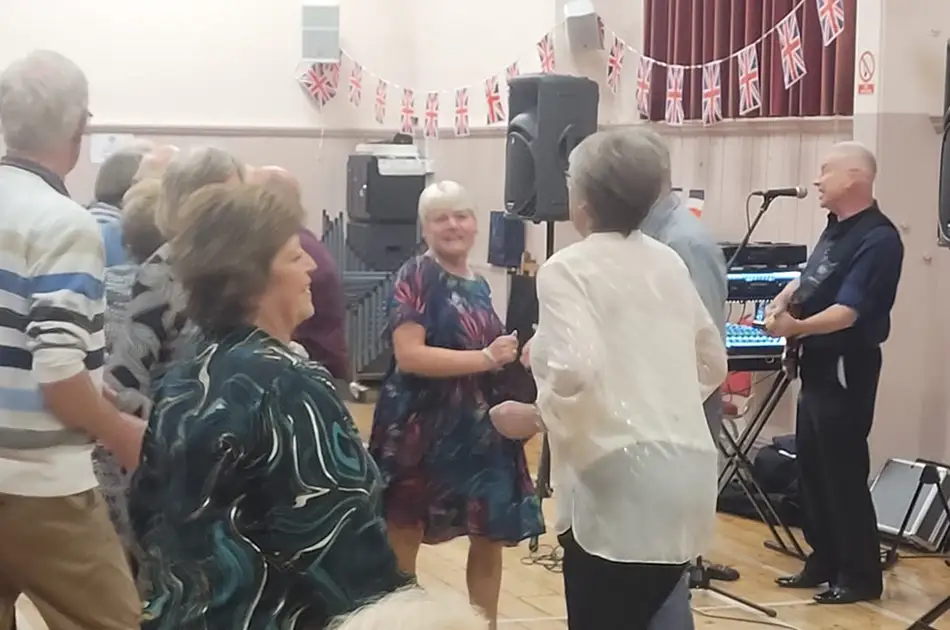 A group of older adults dance and enjoy live music at a Saturday Musical Soirée in a decorated hall with British flag bunting. A man plays keyboard and sings on stage whilst people smile and socialise on the dance floor.