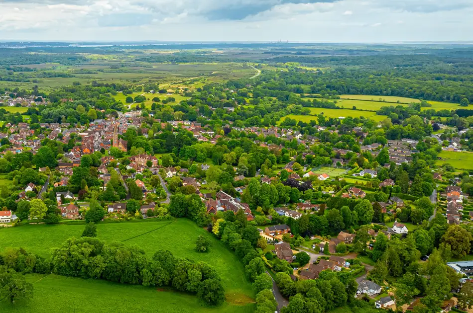 Aerial view of a rural village with houses surrounded by lush green trees and fields, under a cloudy sky, with open countryside stretching into the distance.
