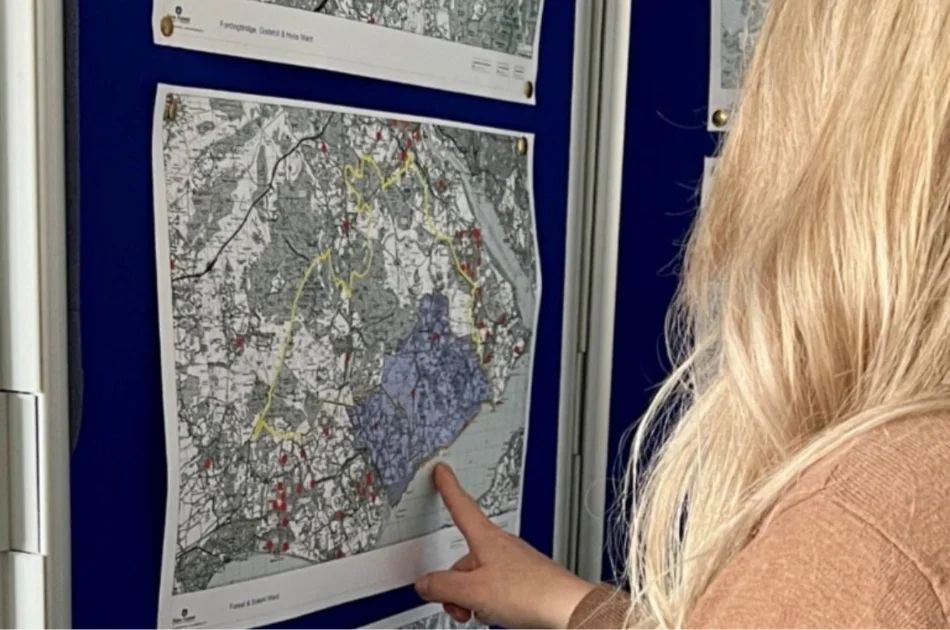 A person with long blonde hair points at a section on a detailed black and white map displayed on a blue board, with other maps visible in the background.